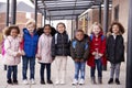 A group of smiling young multi-ethnic school kids wearing coats and carrying schoolbags standing in a row in walkway outside their