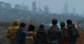 A group of survivors huddle together backs facing the camera as they look towards the horizon. The ruins behind them