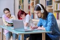 Group of teenage students study sitting at desk in library, in focus is guy using laptop