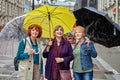 A group of three mature European women stand under umbrellas on a city street in rainy weather.