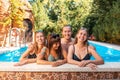 A group of three young women and one man, laughing and posing, leaning on the edge of the pool.Vegetation is in the background.