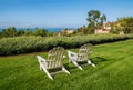 Group of two white Adirondack chairs on green lawn facing a hedge and ocean