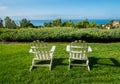 Group of two white Adirondack chairs on green lawn facing a hedge and ocean