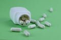 A group of white capsules of medicine spilled out of a white jar on a green background.