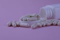 A group of white capsules of medicine spilled out of a white jar on a pink background.