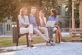 Group of young caucasian adults laughing at bench in a park with their cell phones, laptop, tablet. Looking at camera, eye contact Royalty Free Stock Photo