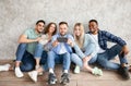 Group of young diverse friends using modern smartphone together, sitting near grey studio wall, full length Royalty Free Stock Photo