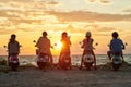 Group of young friends, motorcycle riders wearing helmets standing with their scooters on the coast, enjoying sunset