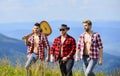Group of young people in checkered shirts walking together on top of mountain. Friendly guys with guitar hiking on sunny