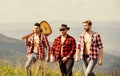 Group of young people in checkered shirts walking together on top of mountain. Friendly guys with guitar hiking on sunny