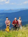 Group of young people in checkered shirts walking together on top of mountain. Men with guitar hiking on sunny day