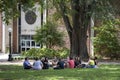 A groups of students sits in a circle on the grass under a tree in front of a law library on a college campus
