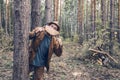 A grown man in a hat and jacket leans out from behind a tree and holds a segment of a log in front of him