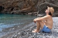 A grown man in a hat sits on pebbles on the seashore