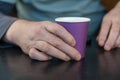 A grown man holds a lilac glass in his hands. A paper cup for hot drinks. Eco-friendly utensils concept. Man sitting at a black