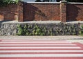 Grunge wall consisting of a base of stones and brick wall above with weeds and ivy. Sidewalk, crosswalk in front.