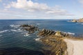 Aerial general view of rocky beach with waves breaking the shore and a couple sitting in the white sand, cliffs and sky with cloud