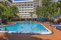 Guests are relaxing on sunbeds under vibrant blue parasols next to central hotel pool, surrounded by lush green palm trees