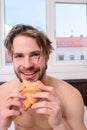 Guy bites sweet dessert. Man bearded handsome guy eating breakfast in bed. Guy holds croissant bedroom hotel room window