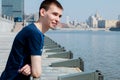 The guy in the blue shirt stands, leaning on the railing on the waterfront of the river, and squinting from the bright sun