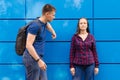 Guy in blue T-shirt and backpack on his back look at girl dressed in blue jeans and plaid shirt against blue wall