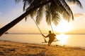 The guy enjoys the sunset riding on a swing on the ptropical beach. Silhouettes of a guy on a swing hanging on a palm tree,