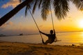 The guy enjoys the sunset riding on a swing on the ptropical beach. Silhouettes of a guy on a swing hanging on a palm tree, Royalty Free Stock Photo