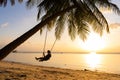 The guy enjoys the sunset riding on a swing on the ptropical beach. Silhouettes of a guy on a swing hanging on a palm tree,