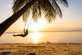 The guy enjoys the sunset riding on a swing on the ptropical beach. Silhouettes of a guy on a swing hanging on a palm tree,