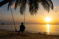 The guy enjoys the sunset riding on a swing on the ptropical beach. Silhouettes of a guy on a swing hanging on a palm tree,