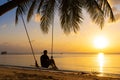 The guy enjoys the sunset riding on a swing on the ptropical beach. Silhouettes of a guy on a swing hanging on a palm tree,