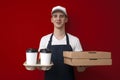 Guy a food delivery uniform in uniform gives coffee and pizza boxes on a red background, food delivery service worker