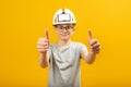 Guy with glasses wears protective helmet and shows thumbs up with two hands. Teenager shows gesture like isolated on yellow