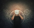 Guy hiding face holding a clock instead of head stands over dark blackboard with drawn gears and cogwheels