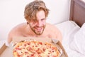 Guy holds pizza box sit bed in bedroom or hotel room. Student is at home on the bed in a bright apartment eating a tasty Royalty Free Stock Photo