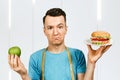 Guy with measuring tape holds a burger and green apple. Concept of choosing between healthy and unhealthy foods Royalty Free Stock Photo