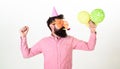 Guy in party hat with air balloons celebrates. Man with beard and mustache on busy face blows into party horn, white