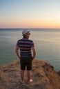 A guy in shorts and straw hat standing on a cliff by the sea