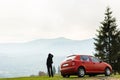 The guy stands near the red car looking at the mountains covered with fog. The sky is pale. Summer. Camping. Picnic. Tourism. Royalty Free Stock Photo