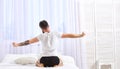 Guy stretching arms, full of energy in morning, rear view. Man in shirt sits on bed, white curtains on background. Macho