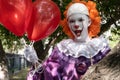 A guy in a IT suit with red balls peeps out from behind a tree. scary clown with scary teeth on the background of an urban fence.