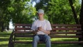 A guy in a sun hat is nervous and fussily looking at the screen of a mobile phone in his hands, while sitting on a bench