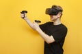 Guy in VR glasses on a yellow background plays a virtual shooter, an emotional gamer shoots a game with joysticks and screams