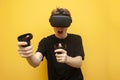 Guy in VR glasses on a yellow background plays a virtual shooter, an emotional gamer shoots a game with joysticks and screams