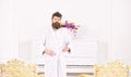 Guy in white bathrobe standing between two antique armchairs. Bearded man tightening his robe. Sleepy man frowning in