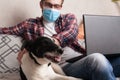 The guy works at home on freelance with his pet. The guy interacts with a dog on the couch with a medical mask