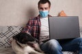 The guy works at home on freelance with his pet. The guy interacts with a dog on the couch with a medical mask Royalty Free Stock Photo