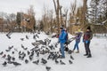 2 guys and a girl in jeans and jackets, with a baby stroller, feeds pigeons in the snowy alley of a city park in winter