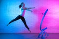 Gymnast woman stretching in front of brick wall in neon lights. Flexible muscular woman doing spirals with ribbon.