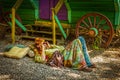 Gypsy man in colorful clothing lays on gravel ground with multi-colored pillows and plays flute by green and purple caravan wagon Royalty Free Stock Photo